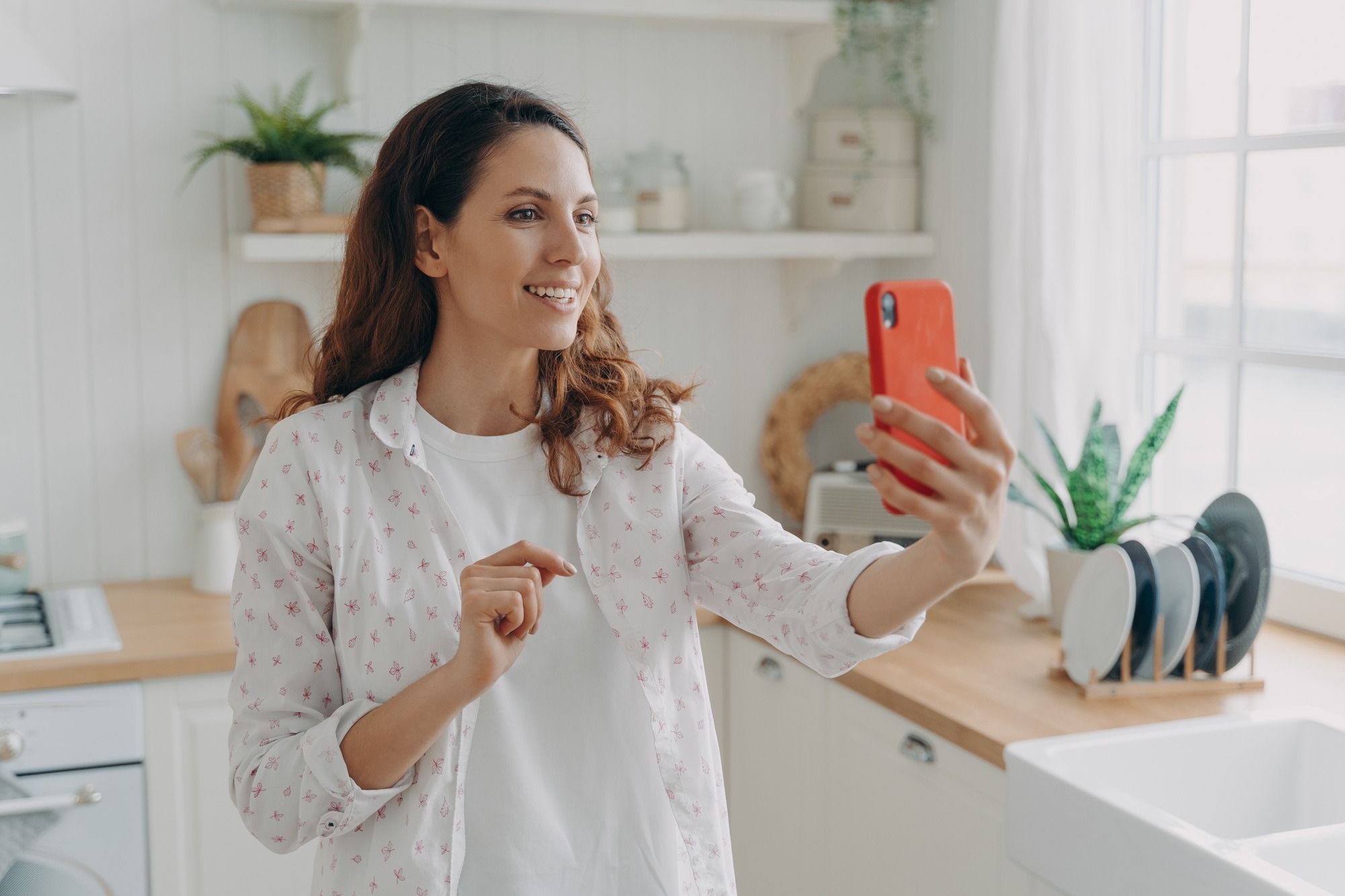Smiling female holding phone, making video call, enjoying online conversation in kitchen at home